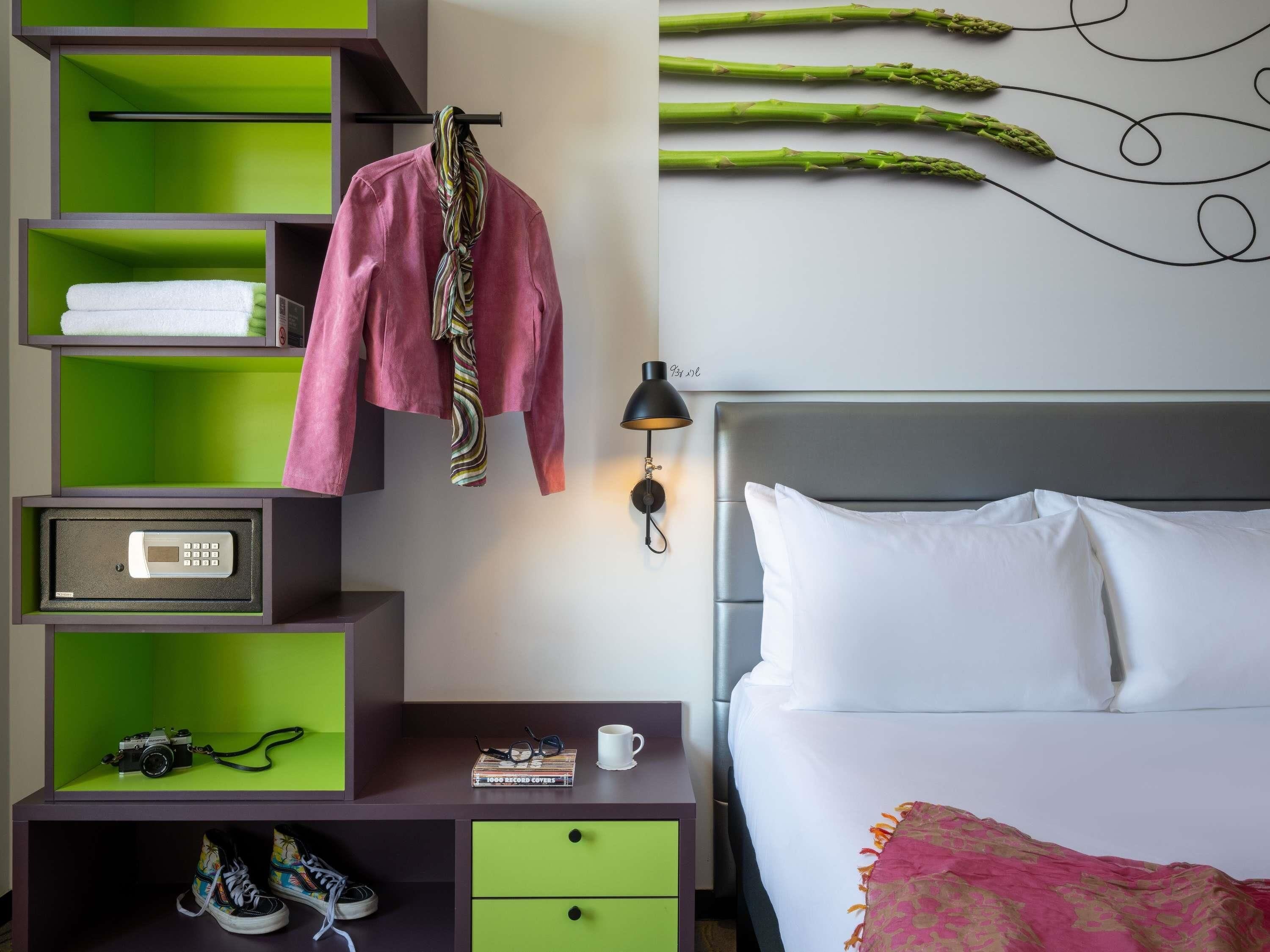 Une chambre d’hôtel avec un lit, une table de chevet, une étagère verte sur laquelle se trouve un appareil photo, une chemise rose et une œuvre d’art représentant des asperges accrochée au mur.