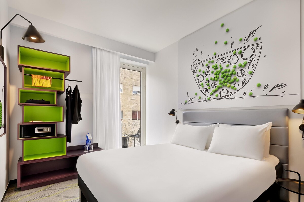 A modern hotel room with a bed, a green shelving unit, a wall art piece, and a view of the outside.