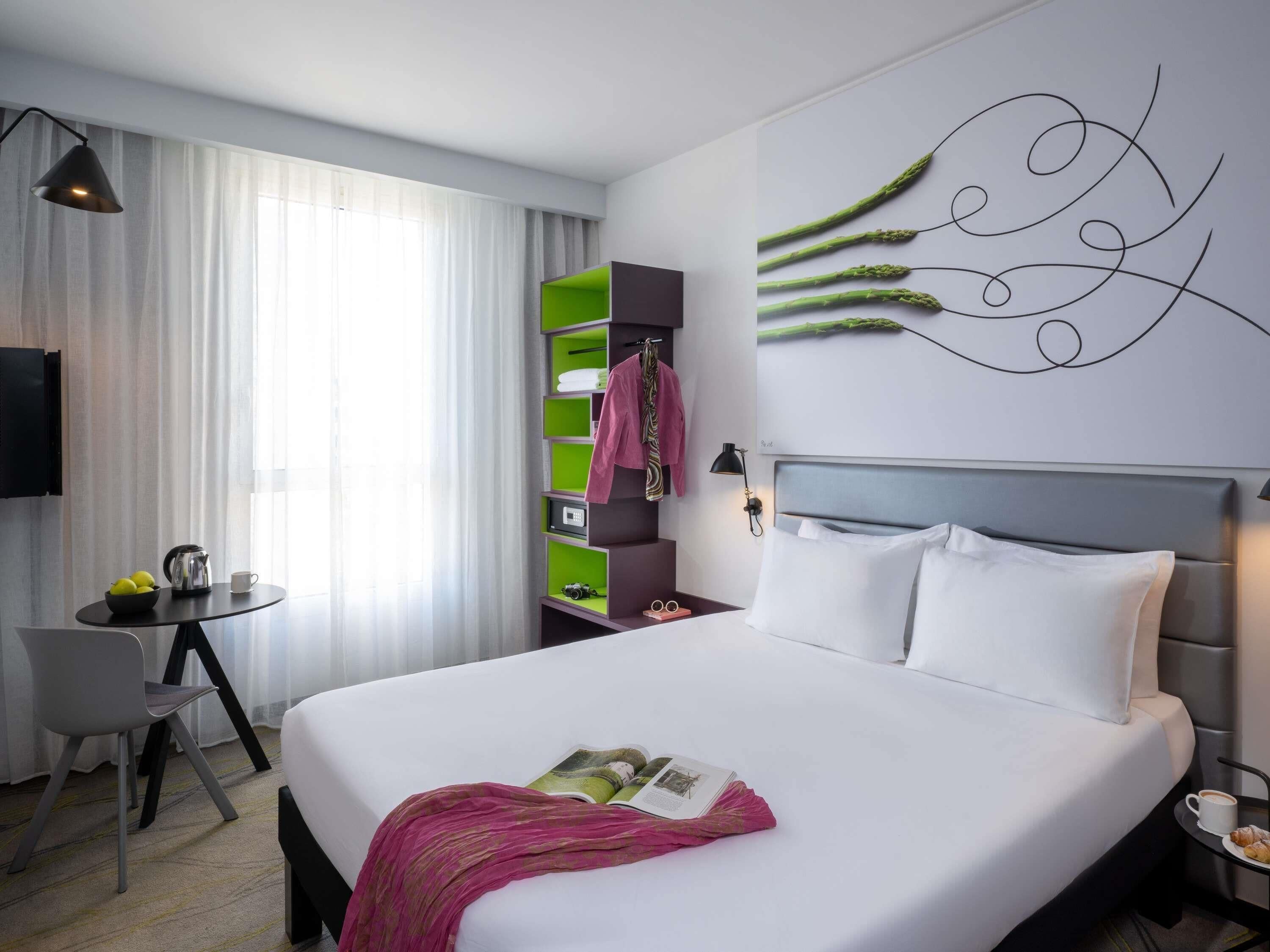 A hotel room with a bed, a desk, a chair, a green storage unit, and a wall art piece.