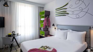 A hotel room with a bed, a desk, a chair, a green storage unit, and a wall art piece.