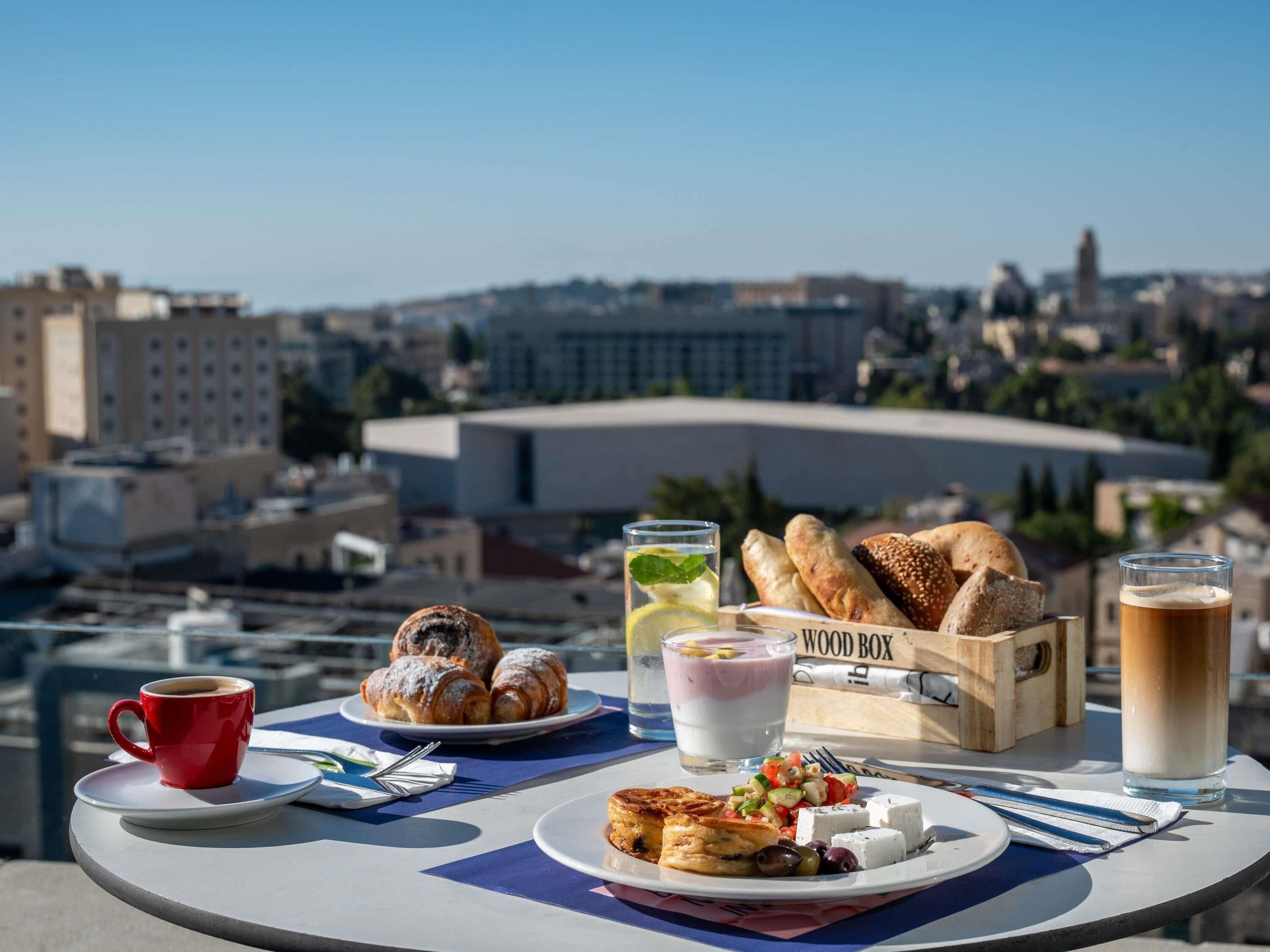 A table with breakfast items including croissants, a cup of coffee, a glass of milk, a bowl of yogurt, and a plate of olives and feta cheese.
