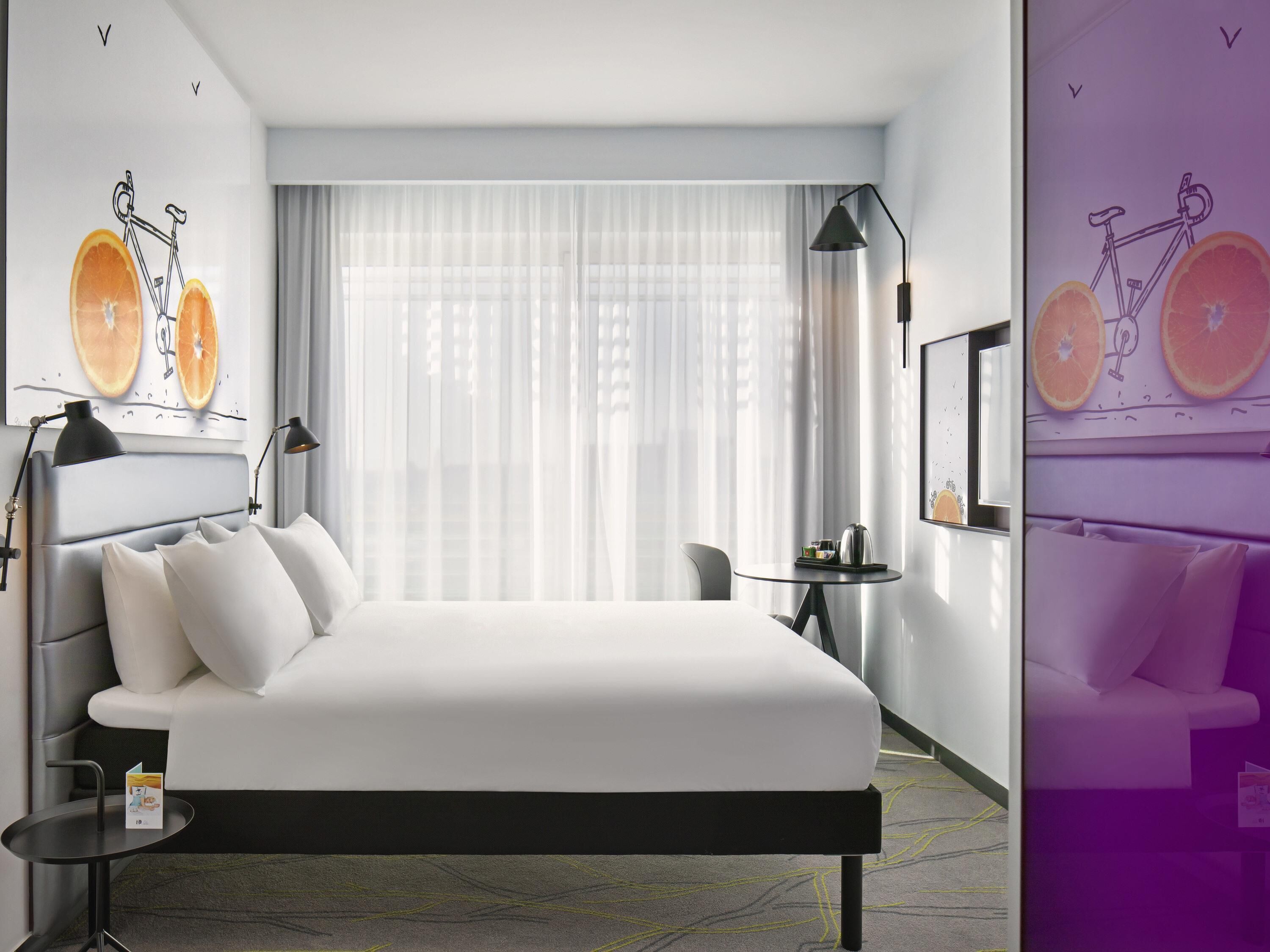 Una camera d'albergo moderna con un letto grande, un tavolino, una sedia e una finestra con tende leggere.