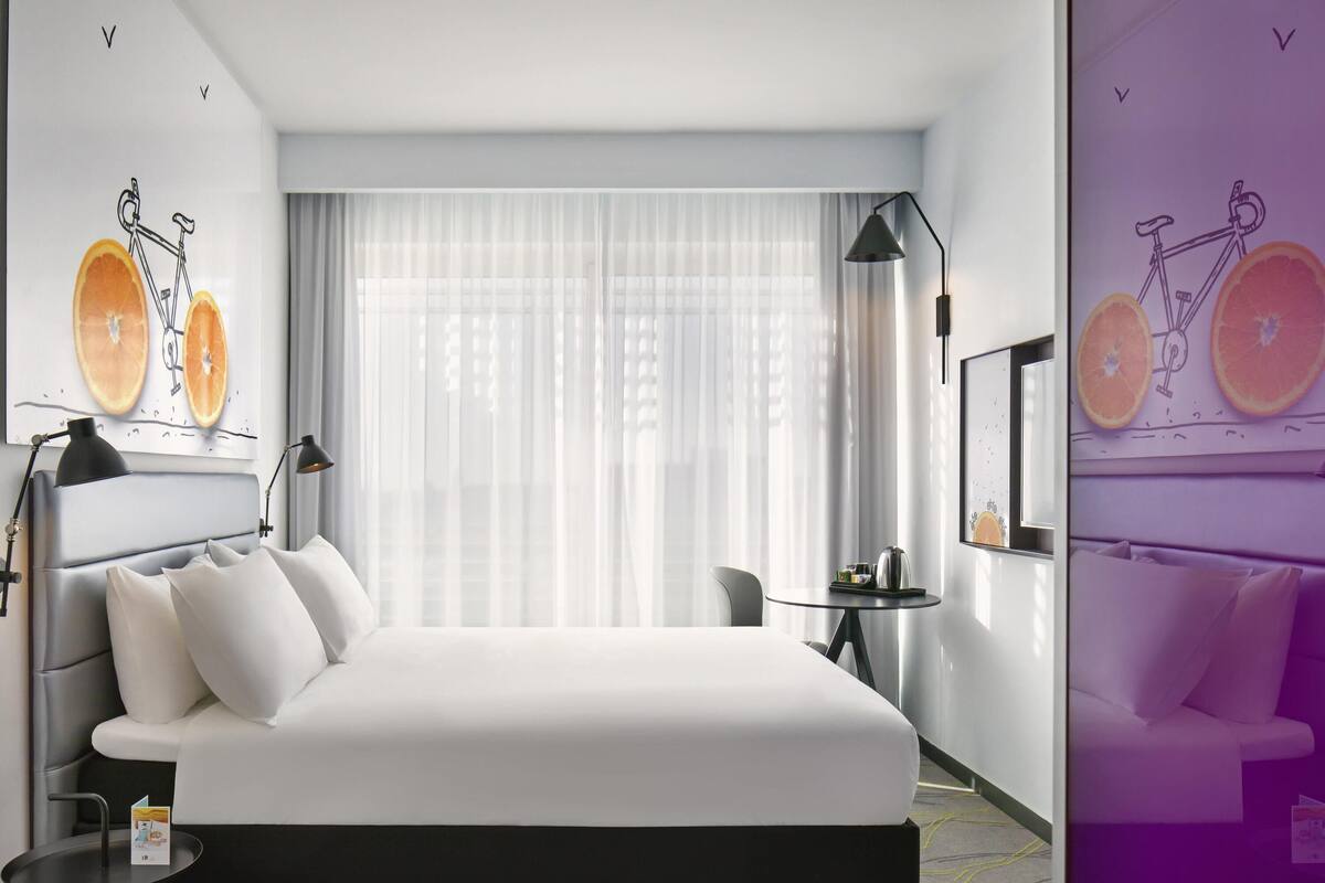 A modern hotel room with a large bed, a small table, a chair, and a window with sheer curtains.