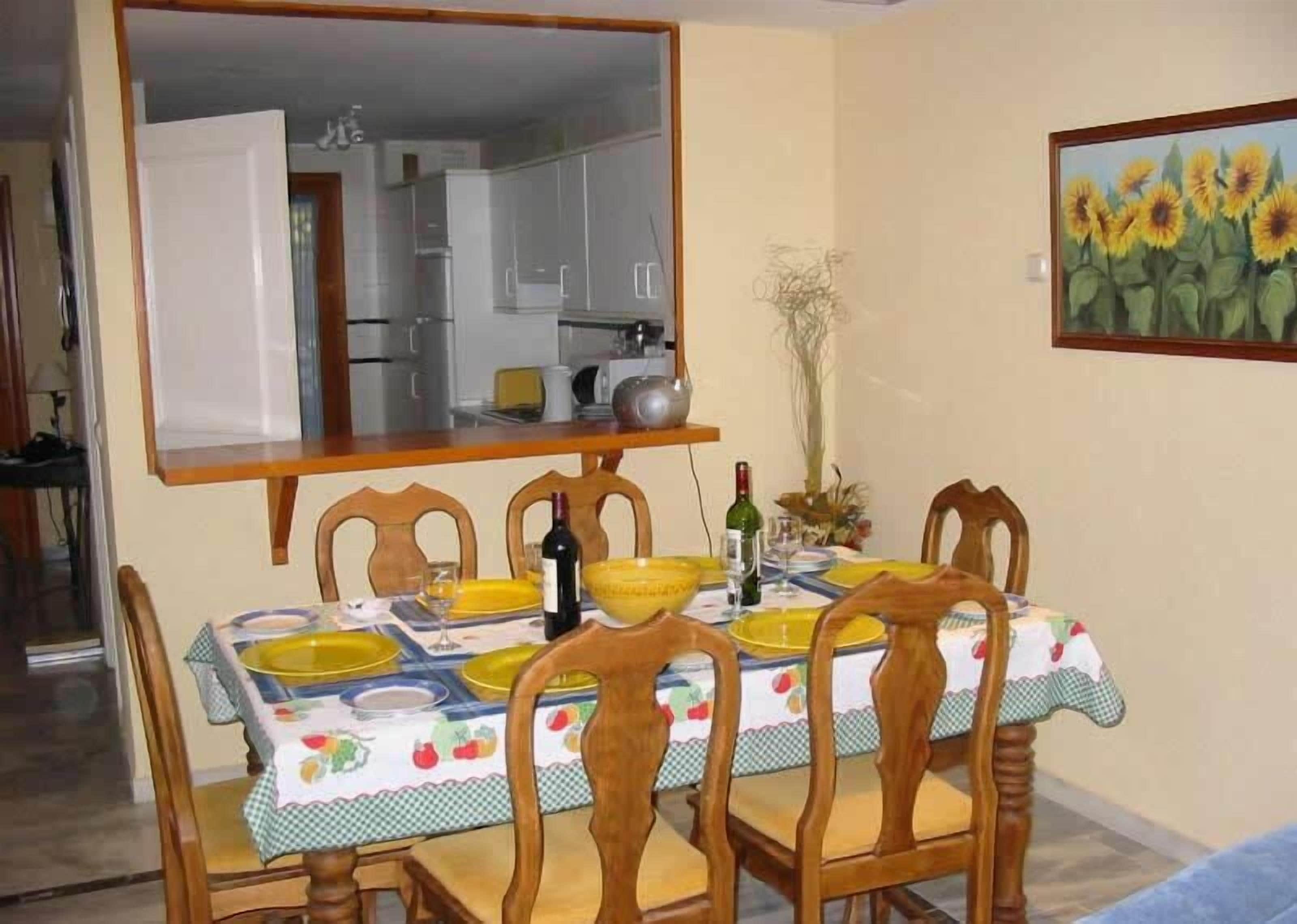 Un coin repas avec une table dressée pour quatre personnes, des chaises, un miroir, un tableau représentant des tournesols et, au fond, une cuisine.