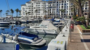 Una marina con varios barcos amarrados, un edificio con balcones y palmeras al fondo.