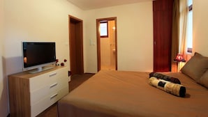 Ein Schlafzimmer mit einem Bett, einem Fernseher auf einer Kommode, einem Fenster mit Vorhängen und einer Tür zu einem Badezimmer.
