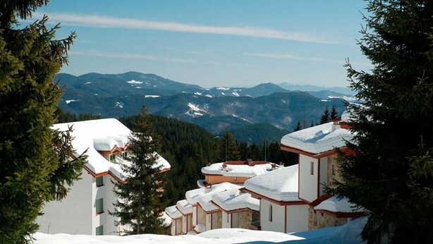 Ein mit Schnee bedecktes Bergresort mit mehreren Gebäuden, darunter eine Hauptlodge und Gästehäuser, umgeben von Kiefern und fernen Bergen.