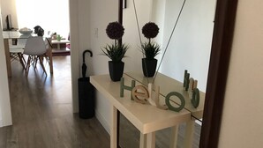 Un couloir avec un plancher en bois, une table blanche ornée de plantes en pot et de lettres en bois formant le mot « Bonjour », un parapluie noir et un miroir accroché au mur.