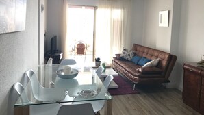 Un salon avec une table à manger en verre, un canapé en cuir marron, un ventilateur de plafond et une vue dégagée sur l’extérieur grâce à une grande fenêtre.