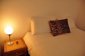 A neatly made bed with white linens and a patterned pillow, a wooden nightstand with a lamp, and a small wooden box.