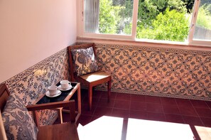 A corner room with patterned walls, a wooden bench, a chair, and a table with two cups and saucers.