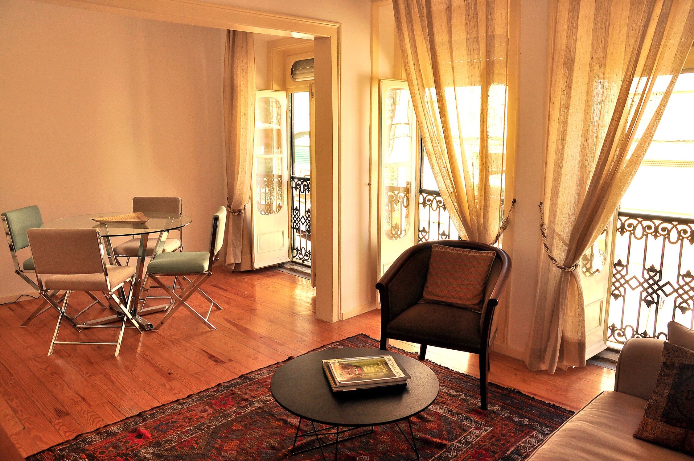 A living room with a glass table, two chairs, a sofa, a patterned rug, and a balcony with curtains.