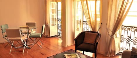 A living room with a glass table, two chairs, a sofa, a patterned rug, and a balcony with curtains.