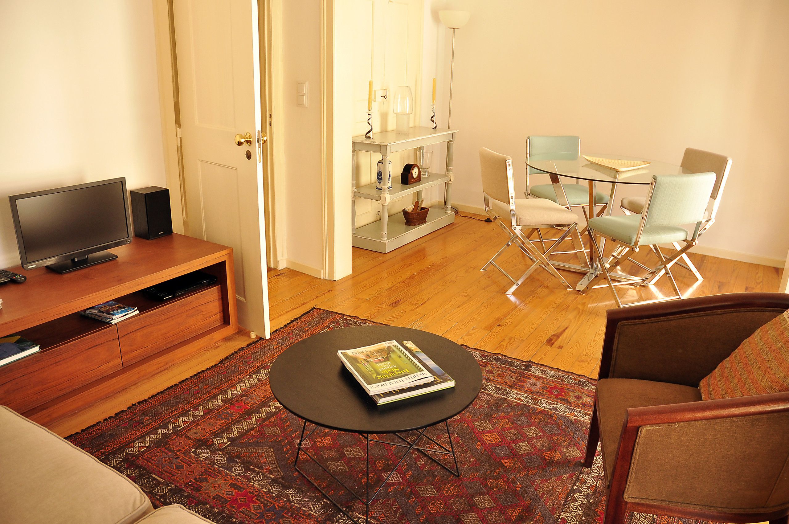 Ein Wohnzimmer mit Fernseher, Couchtisch mit Buch, Essbereich mit Stühlen und Tisch sowie Bücherregal.