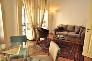 Ein Wohnzimmer mit Glastisch, zwei Stühlen, einer Couch mit Kissen, einer Stehlampe und einem gemusterten Teppich.