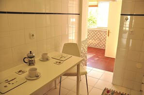 A small dining area with a white table, a chair, and a coffee maker. There is a window with blinds and a door leading to another room.