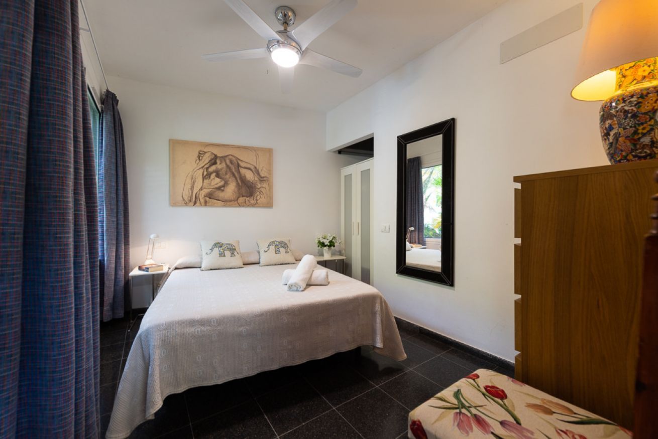 Quarto de hotel com cama, ventoinha no teto, pintura, espelho e guarda-roupa de madeira.