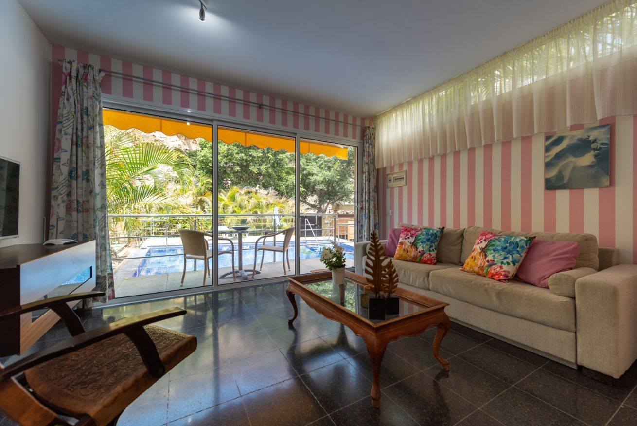 A living room with a sofa, coffee table, and a TV. There is a sliding glass door leading to a balcony with outdoor furniture.