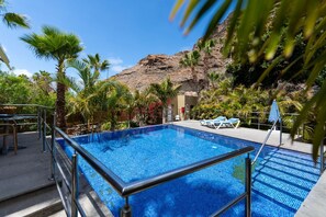 A swimming pool with a metal railing, surrounded by palm trees and a mountain backdrop.
