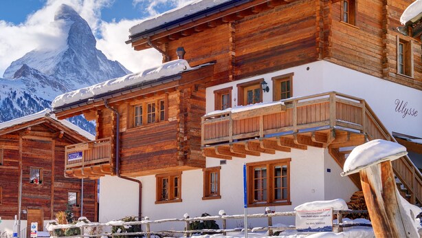 Un chalet en bois doté d’un balcon, une enseigne indiquant « Muttersee Chalets », et une montagne en arrière-plan.