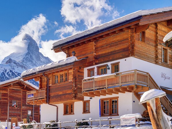 A wooden chalet with a balcony, a sign for 'Muttersee Chalets', and a mountain in the background.