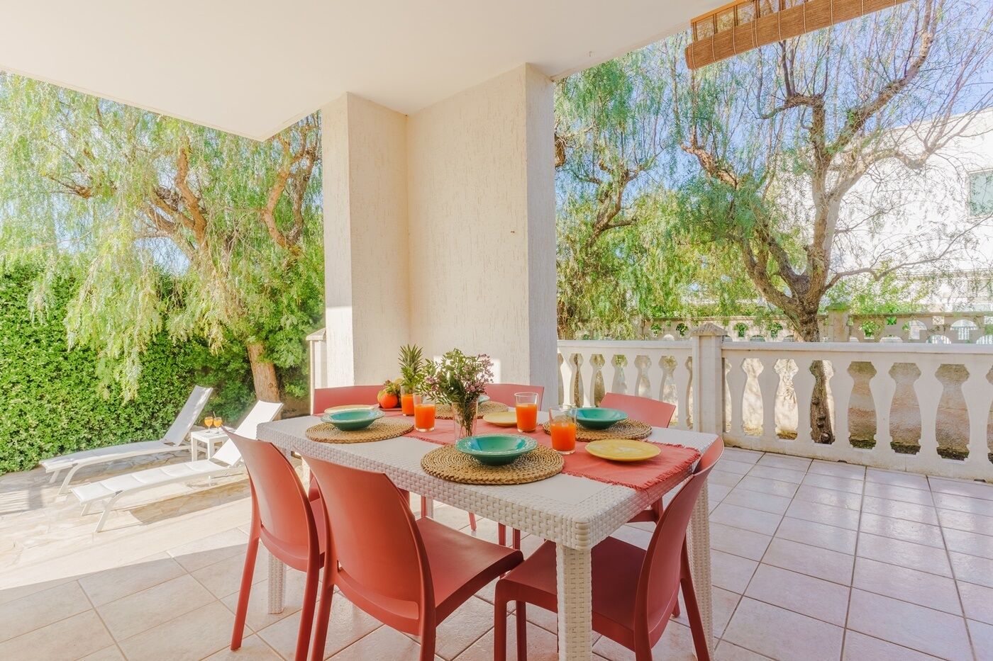 A balcony with a table set for two, surrounded by red chairs, and a view of trees and greenery.