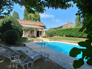 A swimming pool with lounge chairs, surrounded by well-maintained hedges and trees, with a stone building in the background.