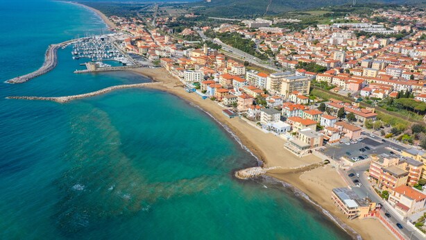 Vista aérea de un pueblo costero con un puerto deportivo, edificios frente a la playa y una red vial clara.