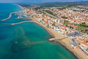 Vista aérea de un pueblo costero con un puerto deportivo, edificios frente a la playa y una red vial clara.