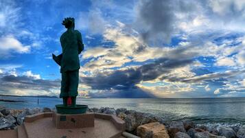 Eine Statue auf einem Sockel am Meer, im Hintergrund ein bewölkter Himmel.