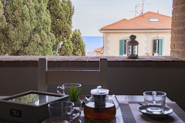 A table with a tea set and glasses, overlooking a building with a tiled roof and a view of the sea.