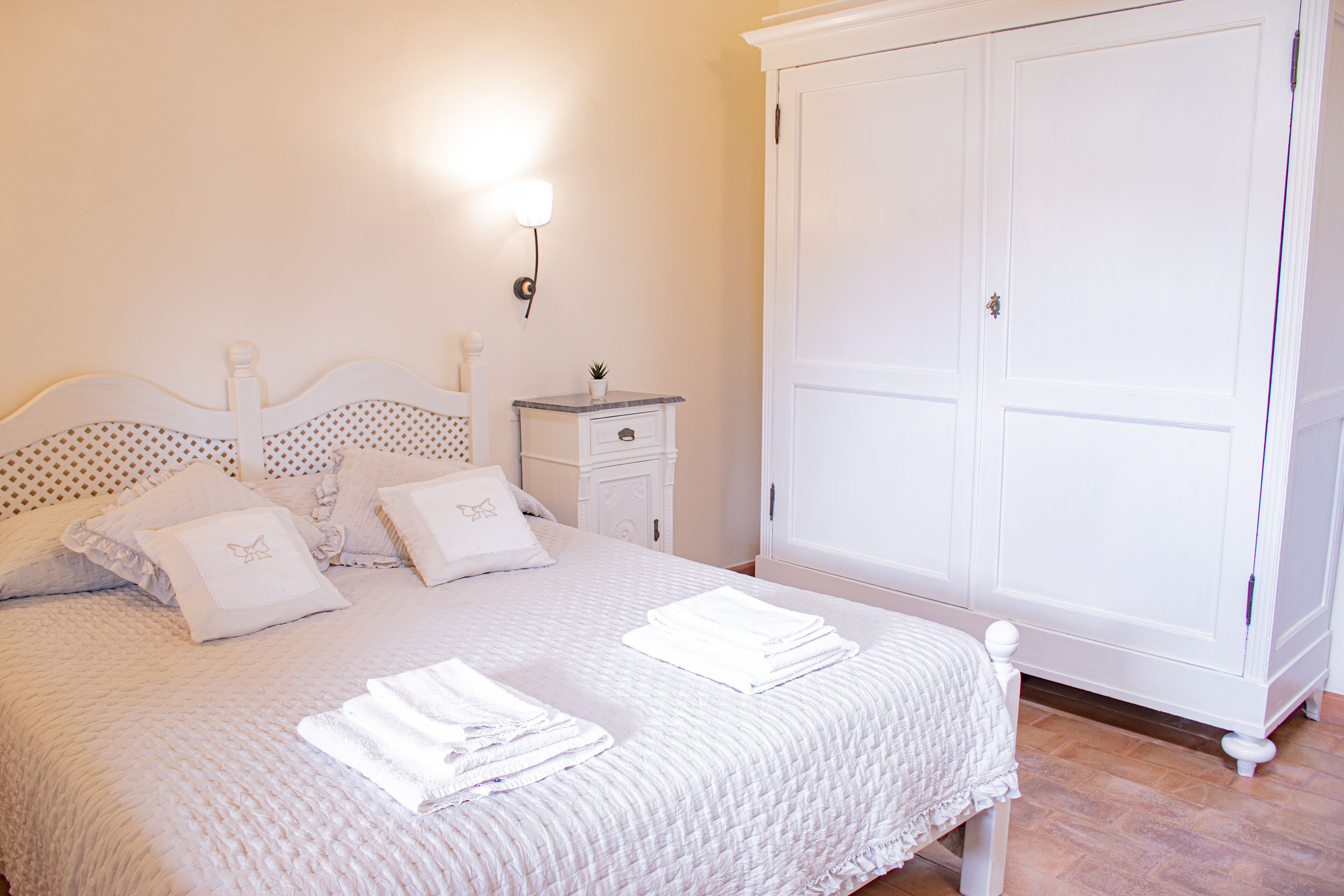 Un lit bien fait, avec une literie et des oreillers blancs, une table de chevet avec une lampe, et une grande armoire blanche.