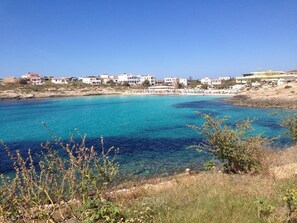 A coastal view with clear turquoise waters, a sandy beach, and buildings in the background. - Villa in Lampedusa for two people,<br>proprietary<br>Nunzia (Lampedusa)