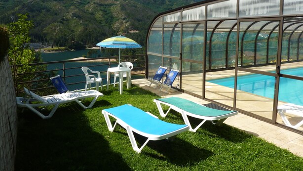 A pool area with lounge chairs and a table, surrounded by a glass enclosure with a view of a lake and mountains.