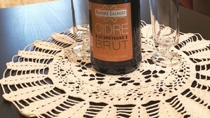 Una botella de Andrée Jalbert Cidre De Bretagne Brut sobre una mesa con un mantel de encaje, vasos y una cocina al fondo.