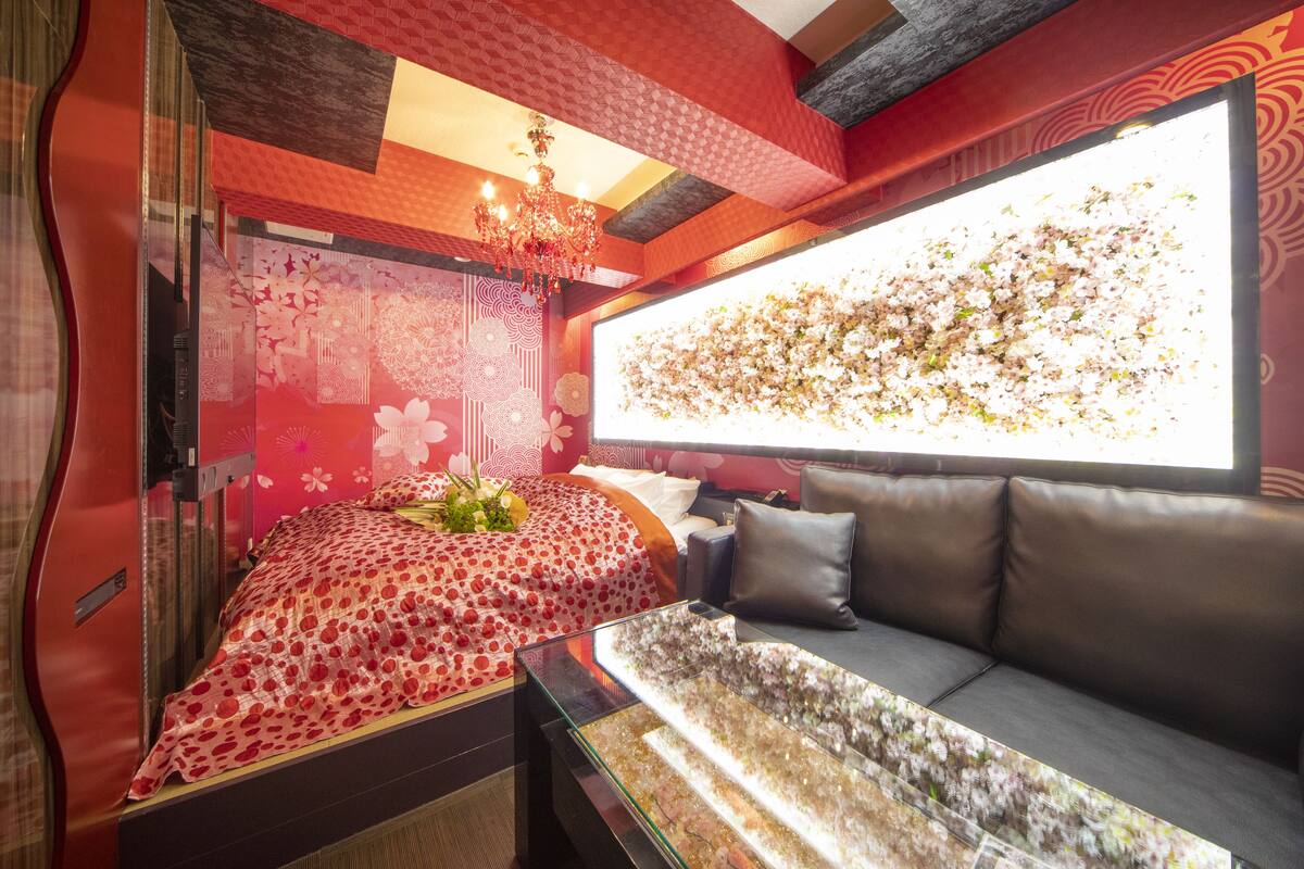 A room with a bed, a sofa, a large window, and a chandelier.