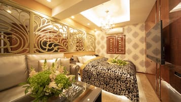A hotel room with a sofa, a bed with a patterned bedspread, a decorative wall panel, a television, and a chandelier.