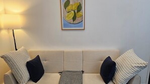 Ein beiges Sofa mit zwei weißen, strukturierten Kissen und zwei dunklen Kissen, einer grauen Decke und einem gerahmten Kunstwerk mit Zitronen an der Wand.