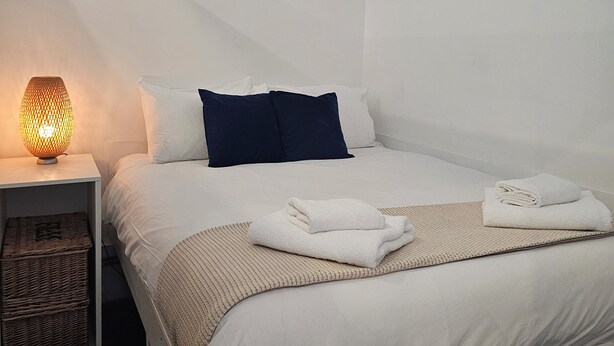 Ein ordentlich bezogenes Bett mit weißen Leinen und einem blauen Kissen, eine Nachttischlampe und ein gefaltetes Handtuch auf einer gewebten Matte.