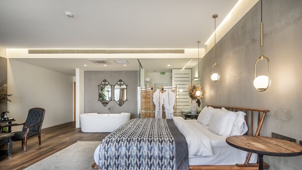 Una habitación de hotel moderna con una cama grande, una mesita de madera, una bañera blanca y una zona de estar con una silla y una mesa.
