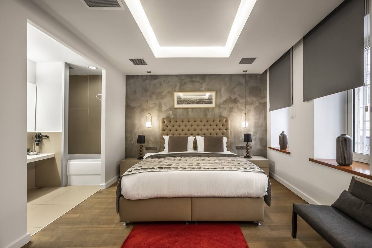 A modern bedroom with a large bed, bedside lamps, a red rug, and a view of the city through the window.