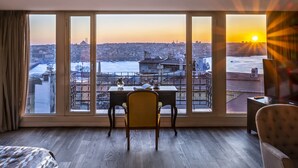 Habitación con un ventanal grande que da a un paisaje urbano y un río. Hay un escritorio con una silla y una mesita con un florero.
