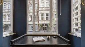 Un escritorio de madera oscura con un libro y una taza, una ventana que da a vistas de un edificio, y una lámpara.