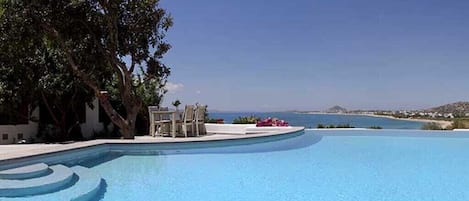 A clear blue swimming pool with a curved edge, surrounded by a white deck and a large tree.
