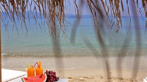 A beach scene with a table under a palm tree, featuring a watermelon slice, grapes, and two glasses of orange juice. - Villa Meadow 5 pax Private Infinity Pool  Beach Hotel Villa Paradise Naxos  (Naxos)