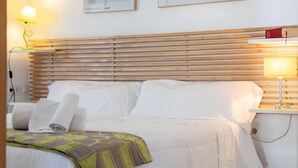 Una cama bien hecha con sábanas blancas, un cabecero de madera y un plaid verde estampado. Hay dos lámparas de noche, una con pantalla amarilla y otra con pantalla blanca.