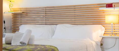 Un letto rifatto con lenzuola bianche, una testiera in legno e una coperta decorata con motivi verdi. Sono presenti due abat-jour, uno con paralume giallo e l'altro con paralume bianco.
