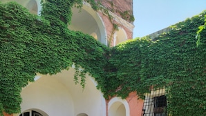 Une cour dallée de pierres, un bâtiment blanc orné d’arcades et une fenêtre avec des barreaux en métal.