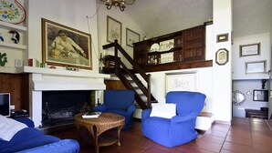 Un salon avec une cheminée, deux fauteuils bleus, une table en osier et un escalier.