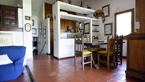 Un salon avec un canapé bleu, une table à manger en bois, des chaises et un coin cuisine doté d’étagères et d’objets décoratifs.